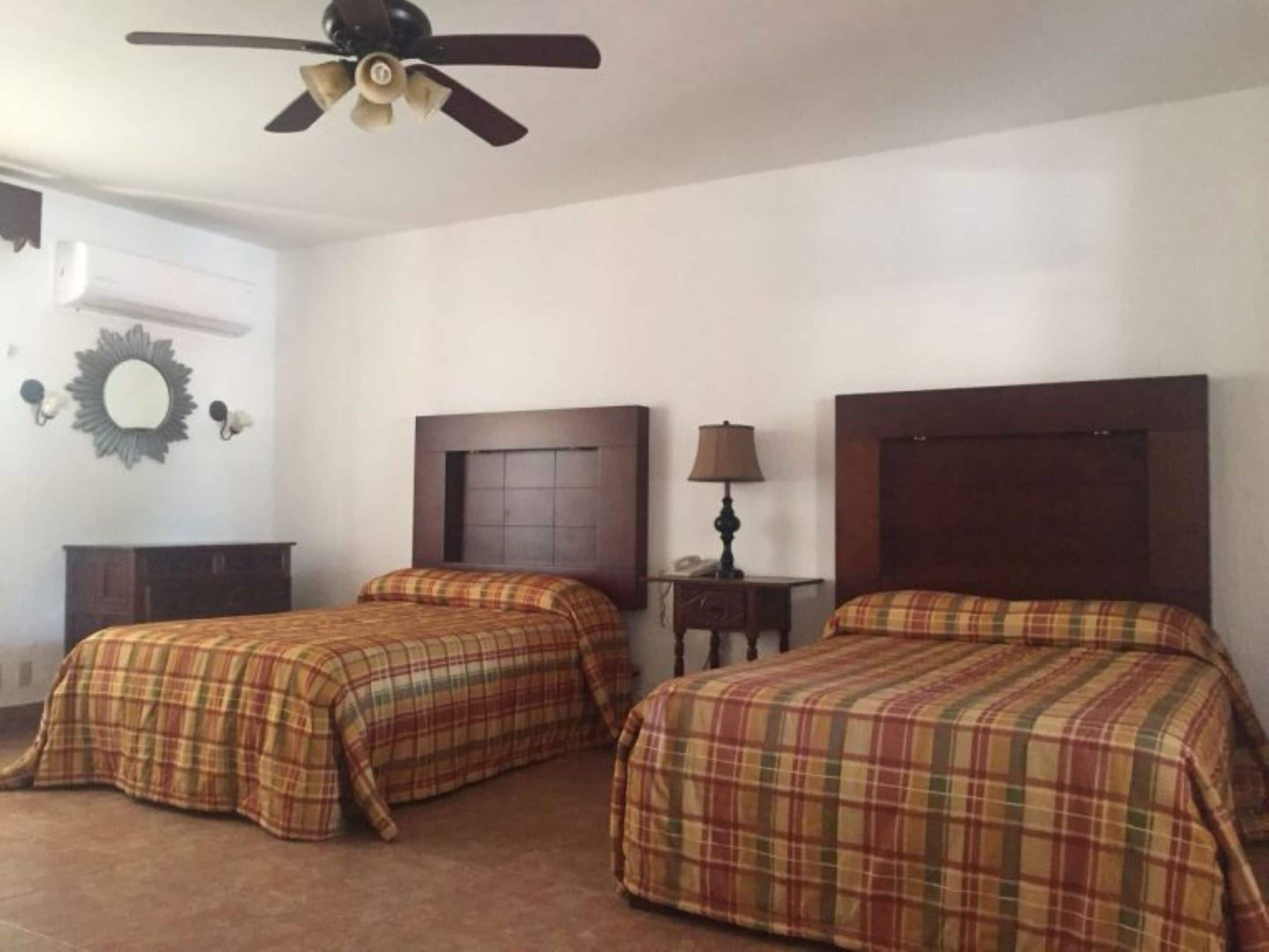 A hotel room with two beds, a ceiling fan, a nightstand with a lamp, and a wall-mounted mirror.