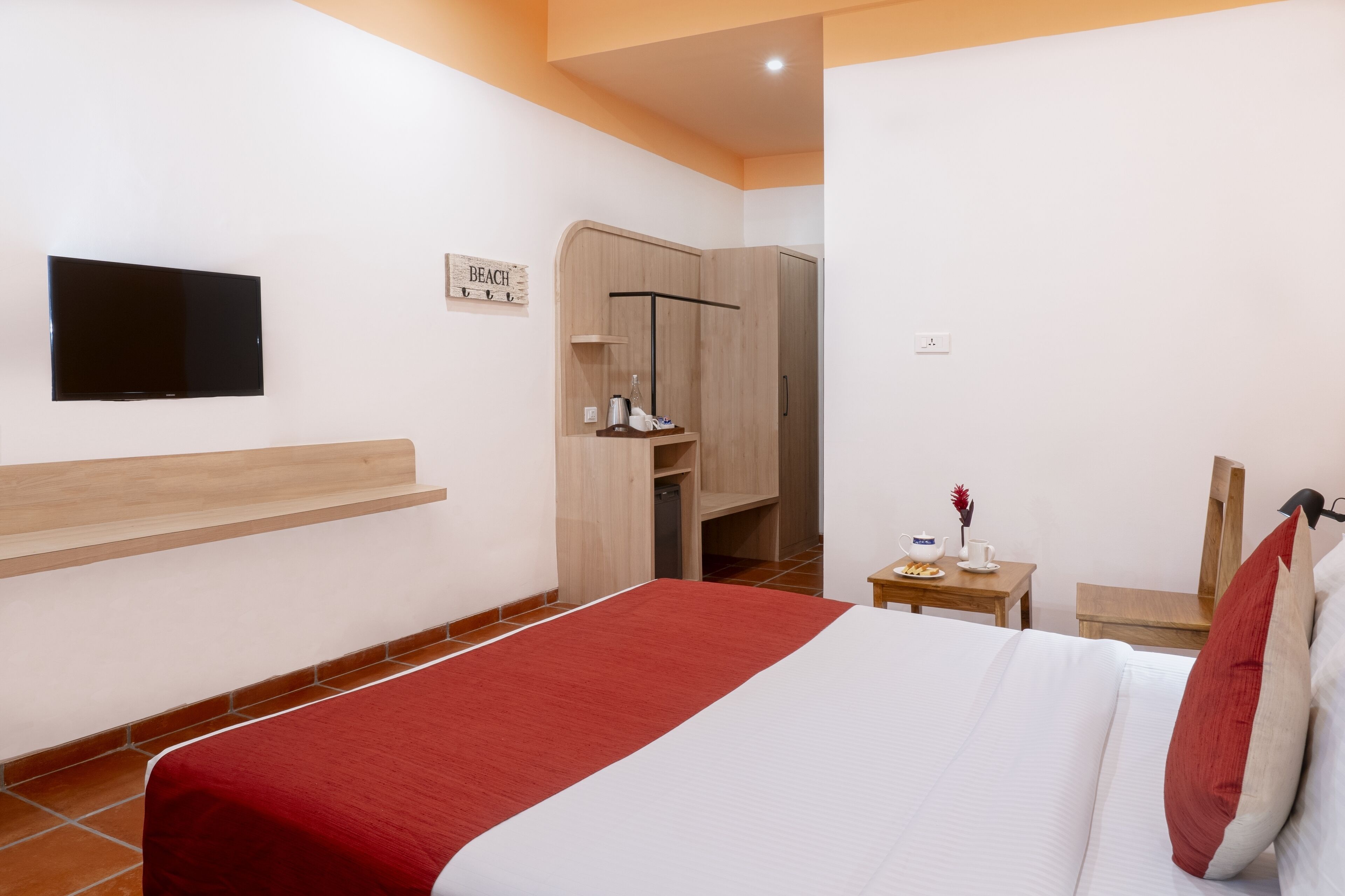 Habitación de hotel con cama, televisor montado en la pared, armario y una mesita de noche con lámpara y florero.