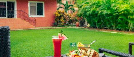 A dining table with a drink, a bowl of fruit, and a sandwich, set against a lush garden backdrop.