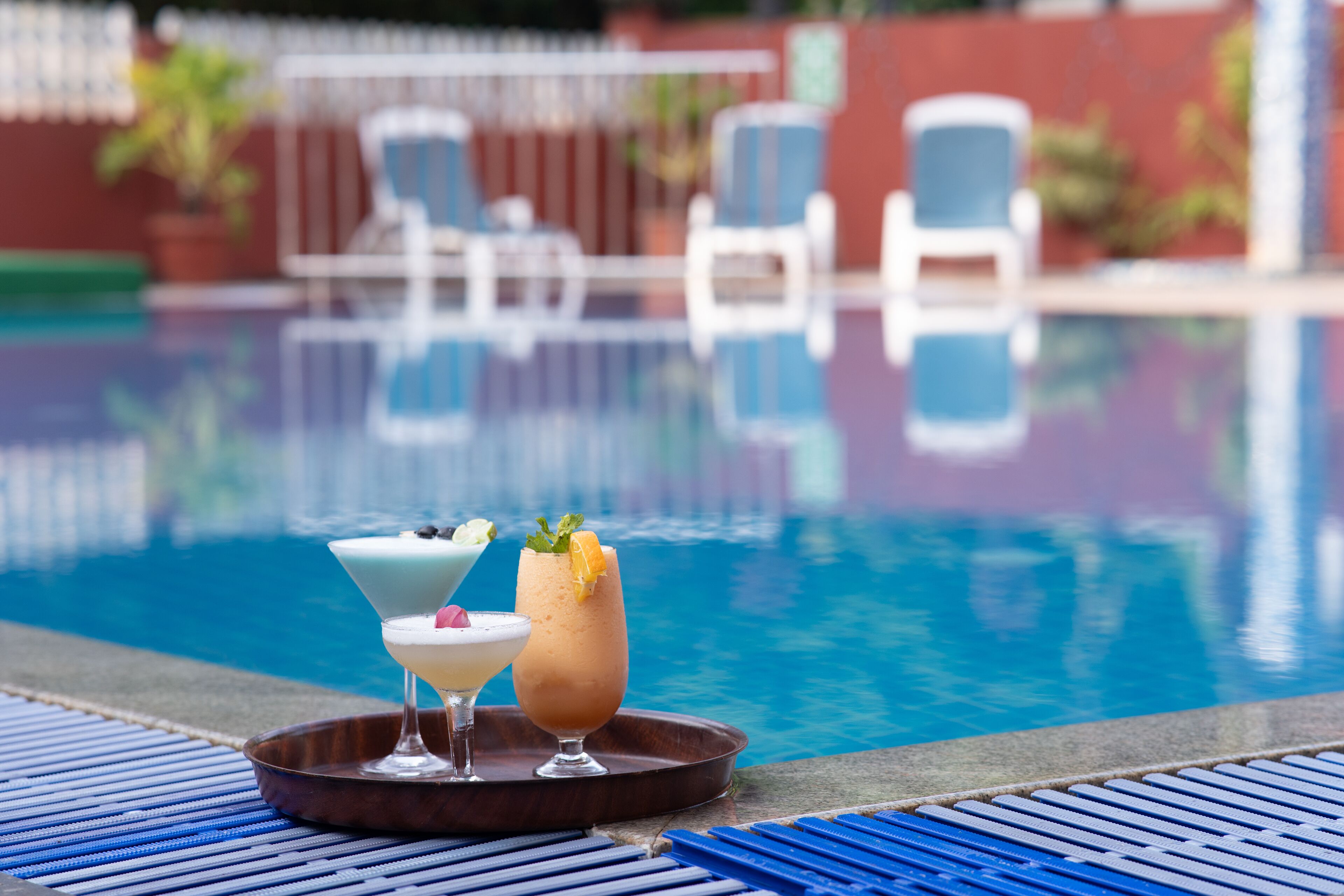 Un bar junto a la piscina con dos cócteles en una bandeja, una piscina al fondo y sillones de descanso.