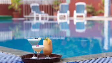 Un bar junto a la piscina con dos cócteles en una bandeja, una piscina al fondo y sillones de descanso.