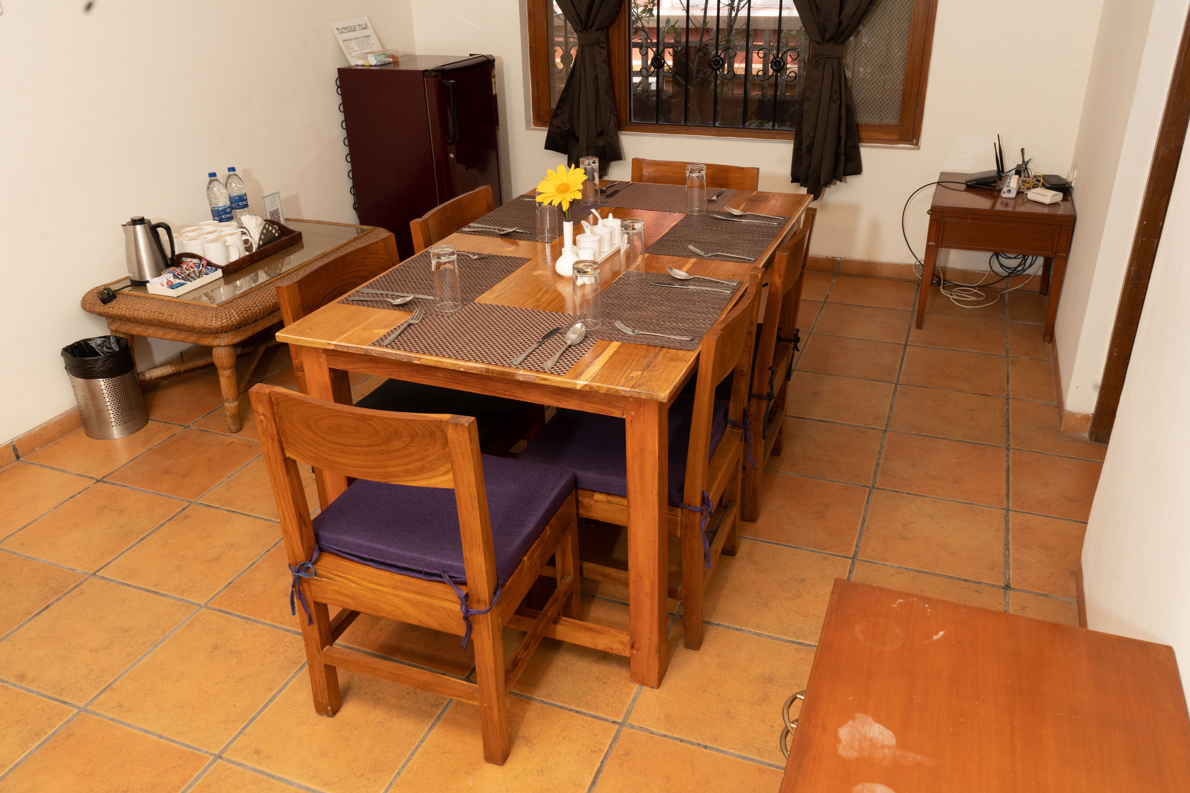 A dining area with a wooden table set for four, surrounded by chairs with purple cushions. A small table with a vase of flowers and a kettle is visible.