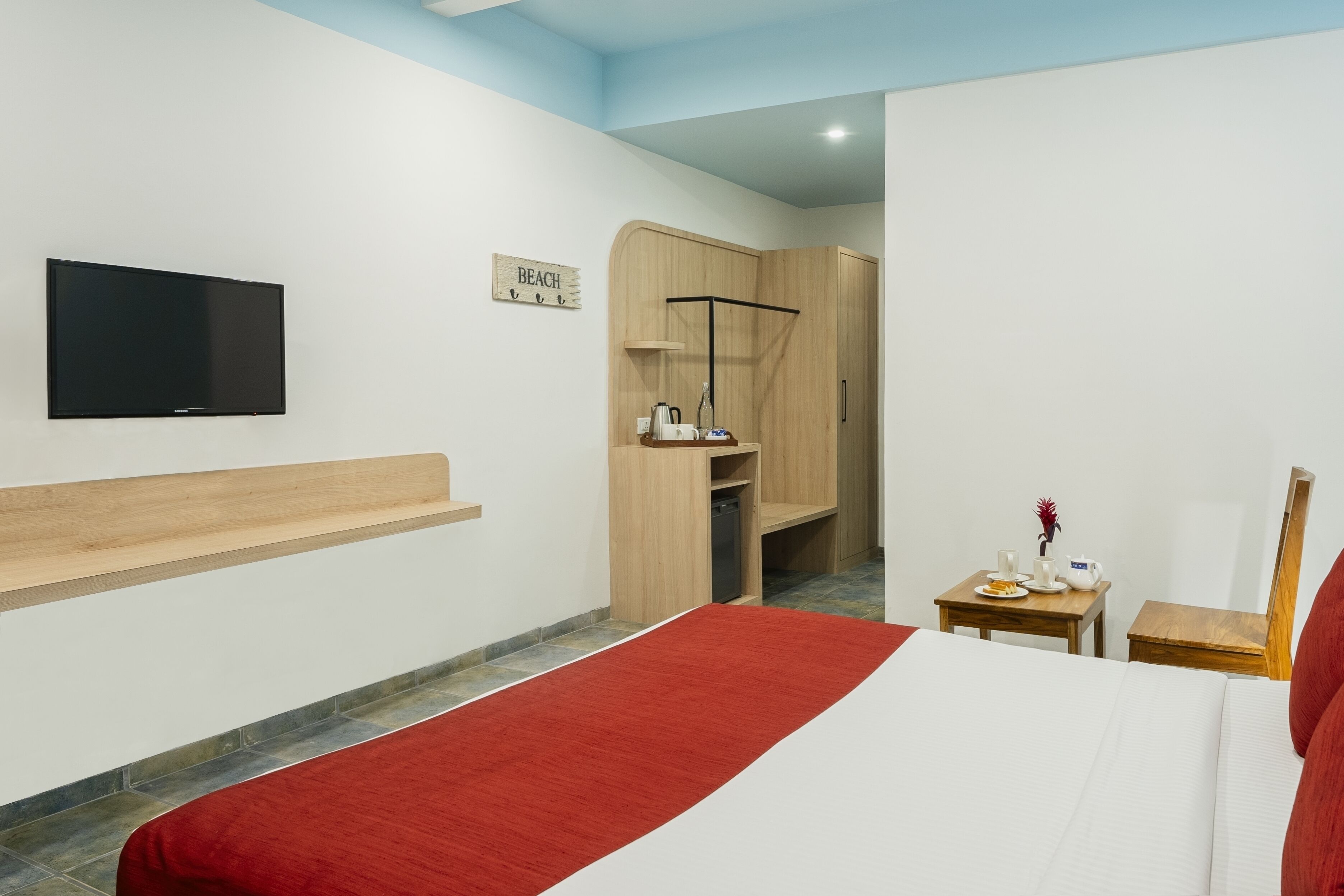 A hotel room with a bed, a TV, a wooden wardrobe, a small table with a cup and a plate, and a sign that says "BEACH".