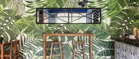 A modern kitchen with a tropical leaf pattern wall, a wooden table with stools, and a bar area with patterned tiles.