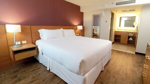 Habitación de hotel con una cama grande, cabecero de madera, mesitas de noche con lámparas, baño con espejo y lavamanos, y piso de madera.