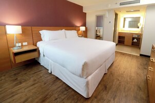 Habitación de hotel con una cama grande, cabecero de madera, mesitas de noche con lámparas, baño con espejo y lavamanos, y piso de madera.