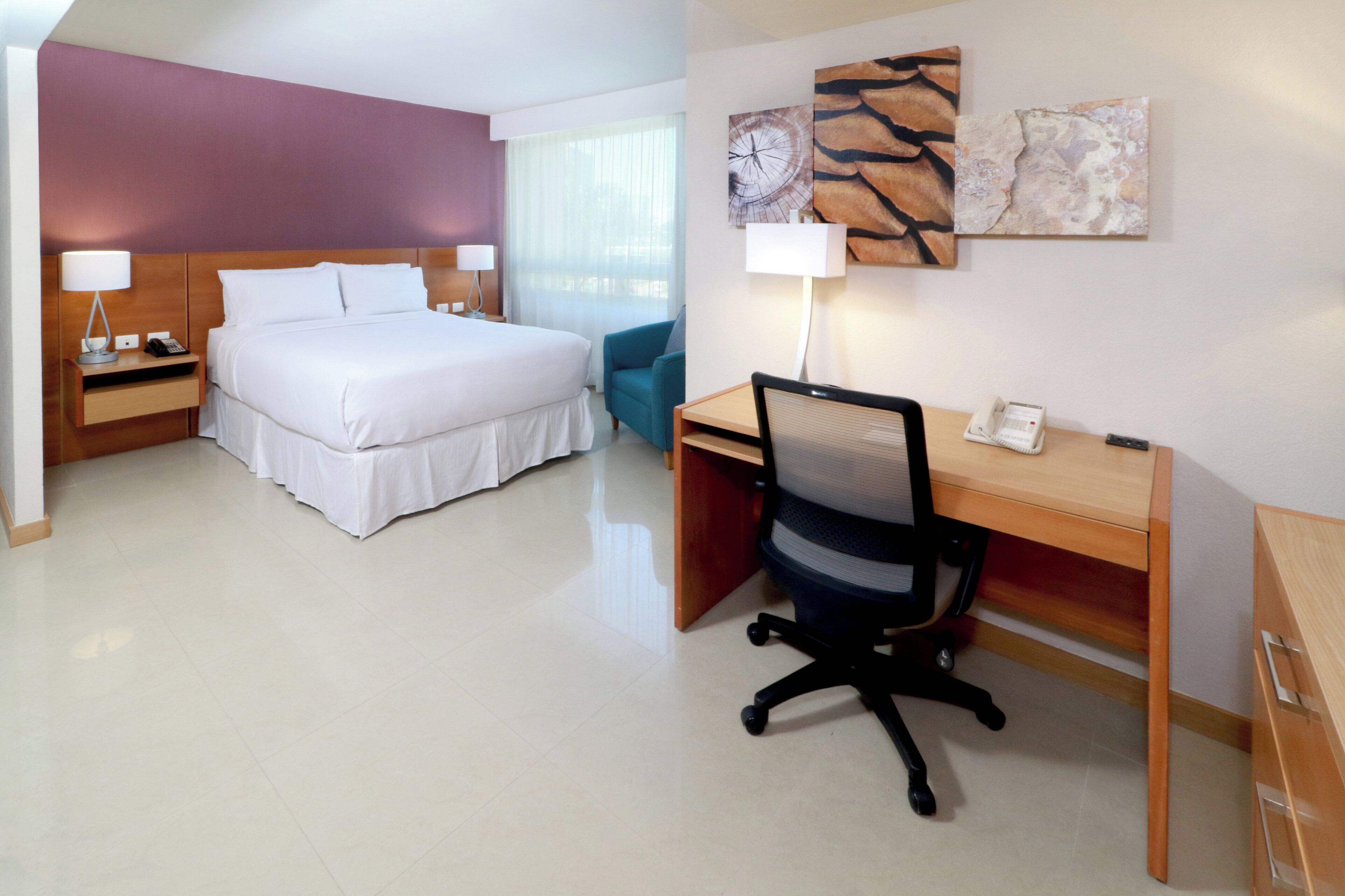 A hotel room with a bed, desk, chair, and artwork on the wall.