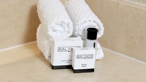 Una esquina de un baño con dos toallas blancas enrolladas, dos cajas etiquetadas 'BALANCE BODY MASSAGE SOAP' y 'BALANCE FACIAL SOAP', y una botella de jabón.