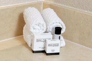 A corner of a bathroom with two white towels rolled up, two boxes labeled 'BALANCE BODY MASSAGE SOAP' and 'BALANCE FACIAL SOAP', and a bottle of soap.