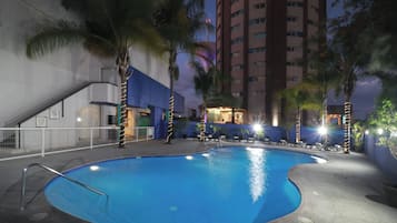 Una piscina de borde curvo, rodeada de palmeras y sillas de descanso, con un rascacielos al fondo.