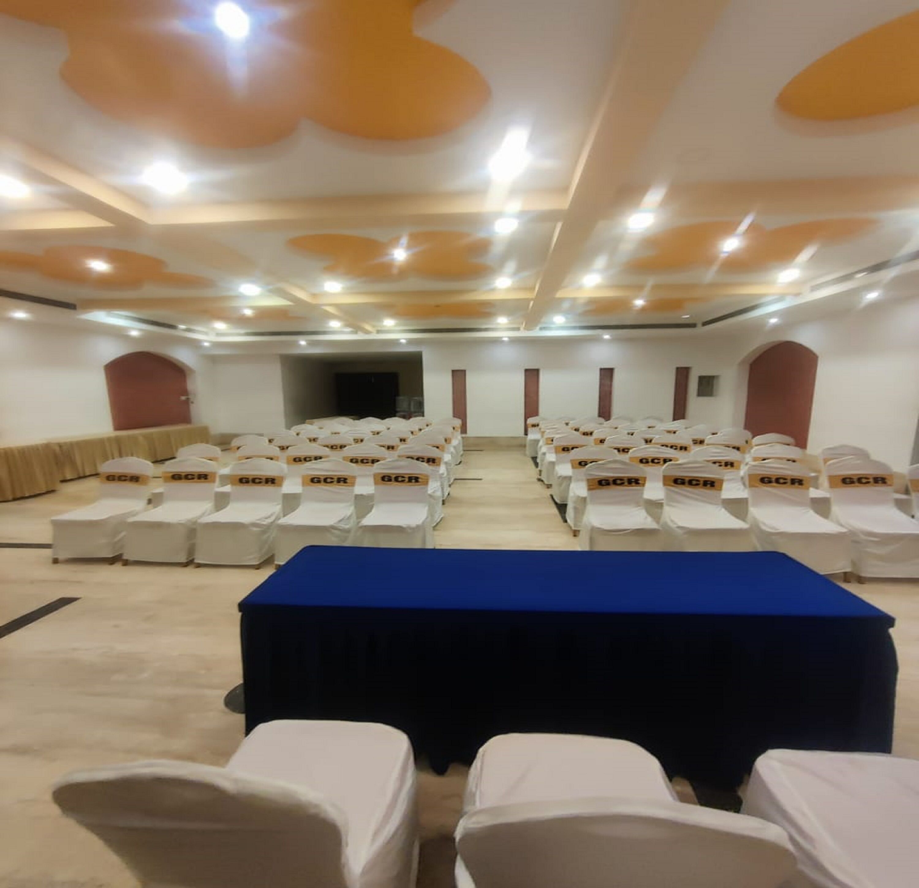 Ein Konferenzraum mit Reihen weißer Stühle, einem blauen Tisch und einer Decke mit eingelassener Beleuchtung.