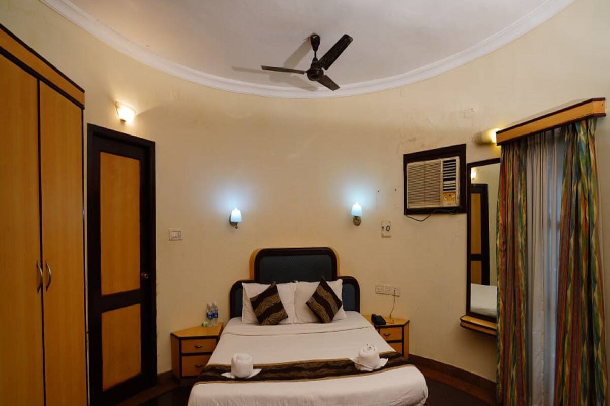 Une chambre d’hôtel comprenant un lit, des tables de chevet, un ventilateur de plafond, des lampes fixées au mur, une fenêtre avec des rideaux et une armoire.