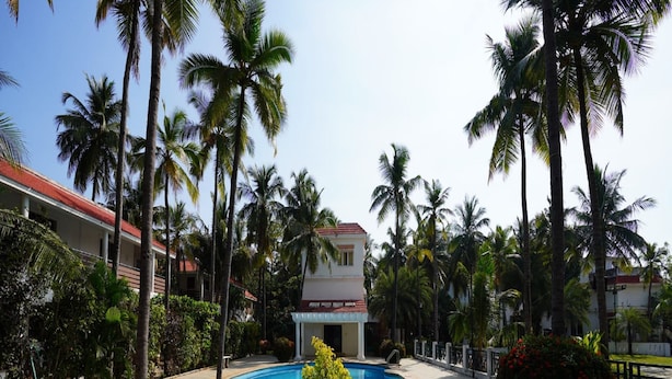 A swimming pool with a circular design, surrounded by palm trees and a building in the background. - Green Coconut Resort (Tirupporur)
