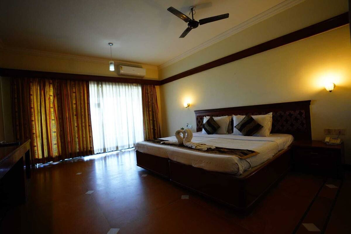 Une chambre d’hôtel spacieuse, dotée d’un grand lit, d’un ventilateur de plafond, d’un appareil de climatisation et offrant une vue sur l’extérieur à travers de fines rideaux.