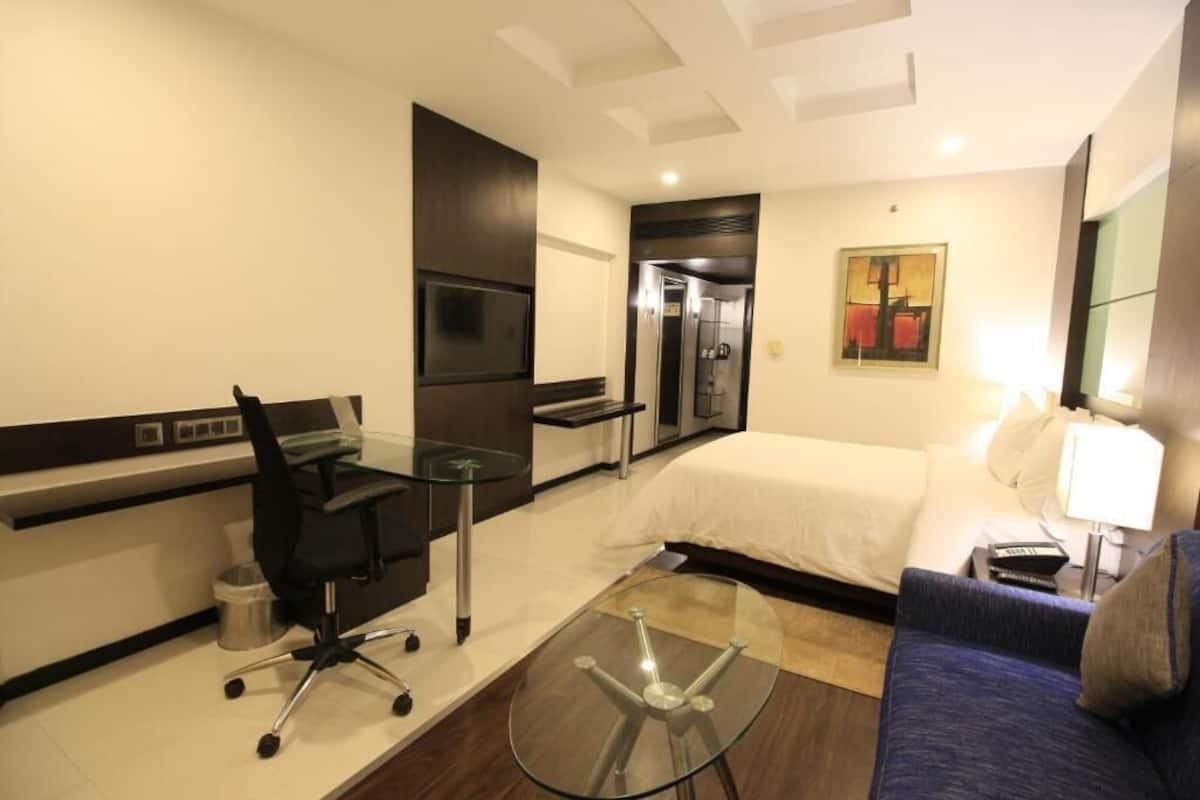 A hotel room with a bed, a desk with a chair, a glass table, a sofa, a TV, and a painting on the wall.