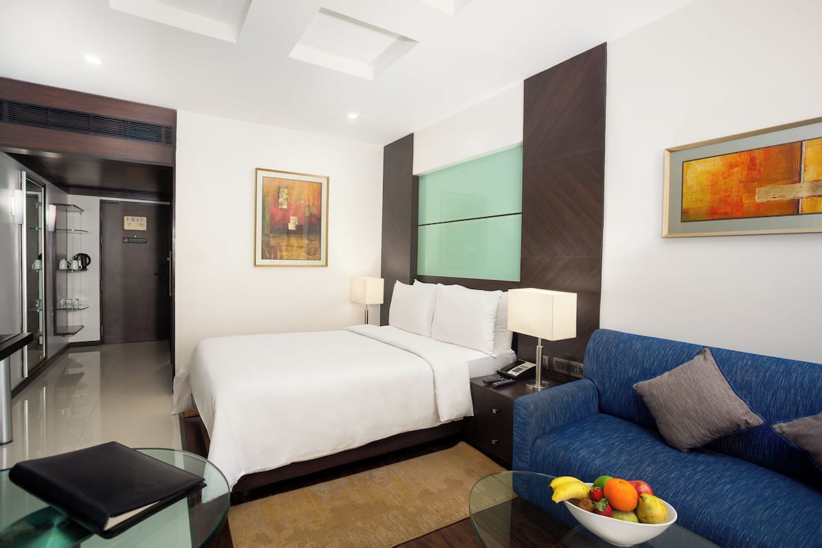 A modern hotel room with a bed, a blue sofa, a glass coffee table, and a fruit bowl.