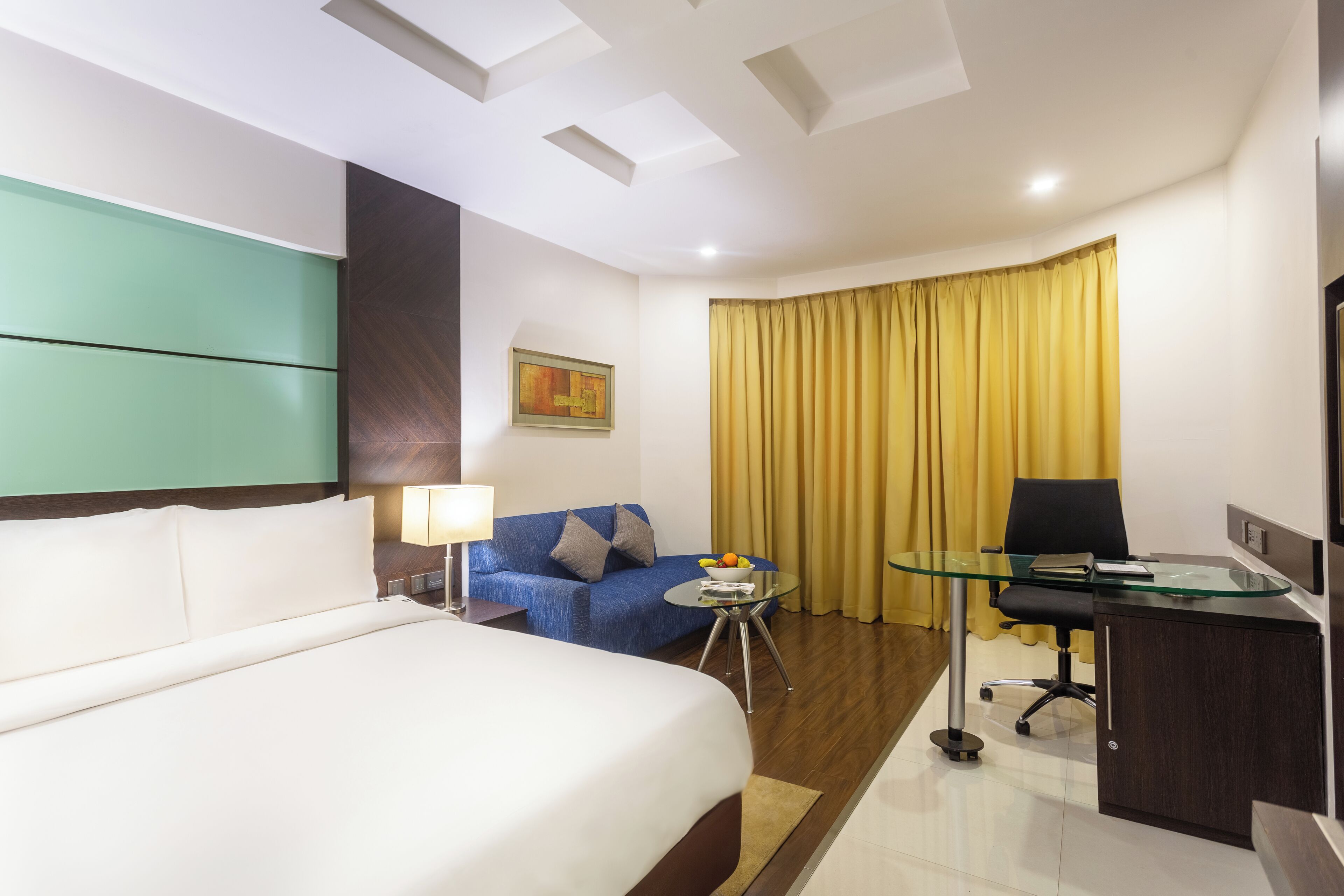 A hotel room with a bed, a sofa, a desk, a glass table, and a yellow curtain.