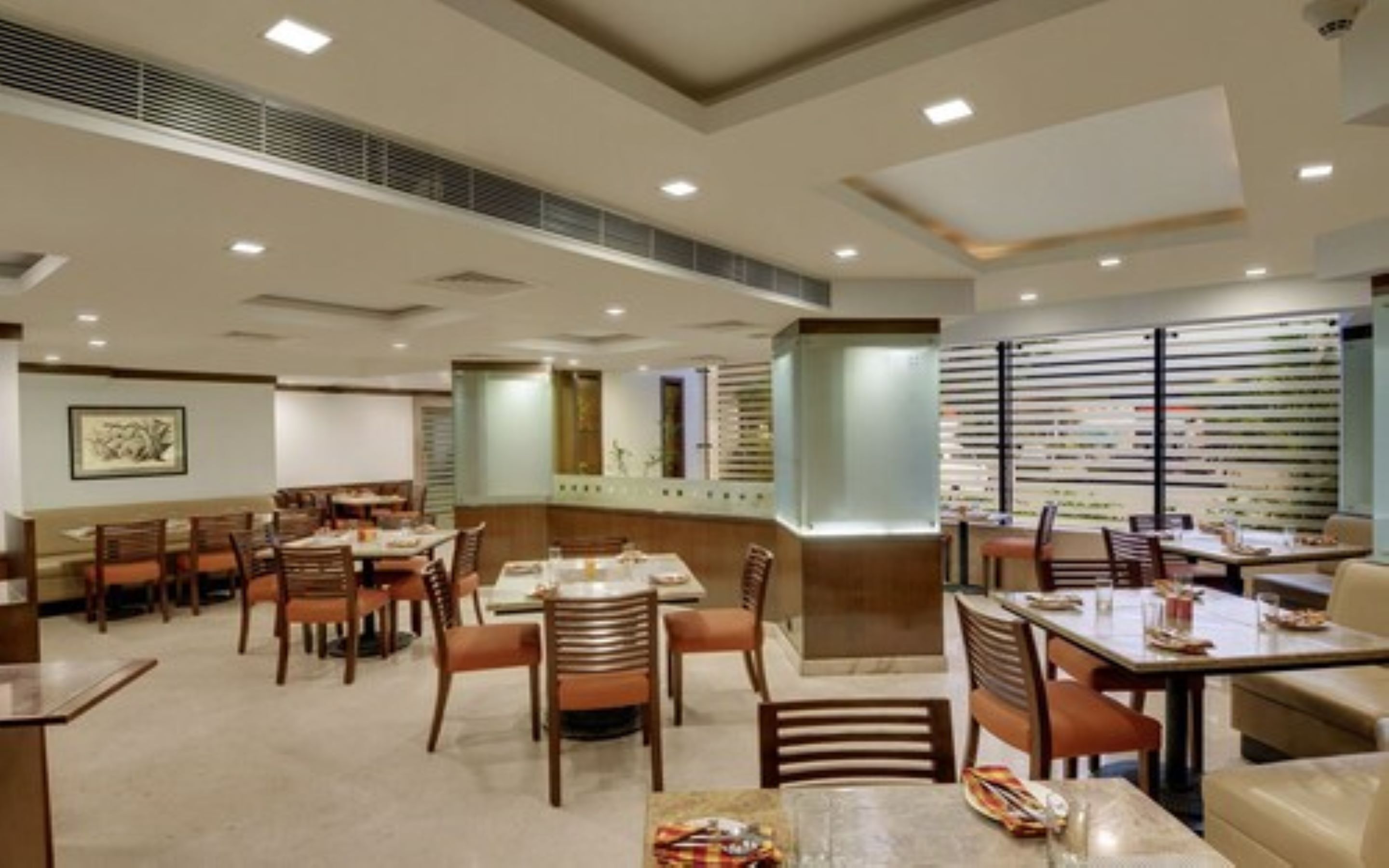 A hotel dining area with multiple tables and chairs, a large window with blinds, and a framed picture on the wall.