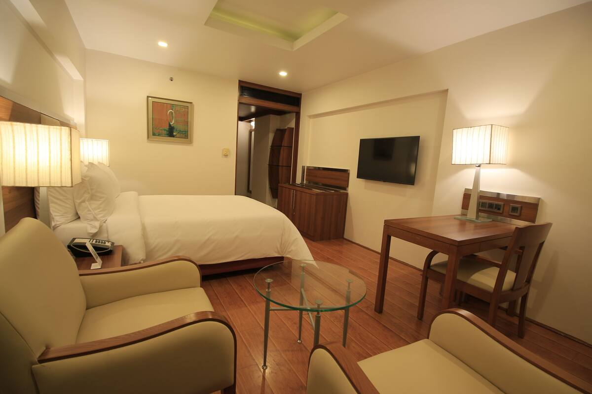 A hotel room with a bed, two armchairs, a glass table, a TV, and a wooden cabinet.