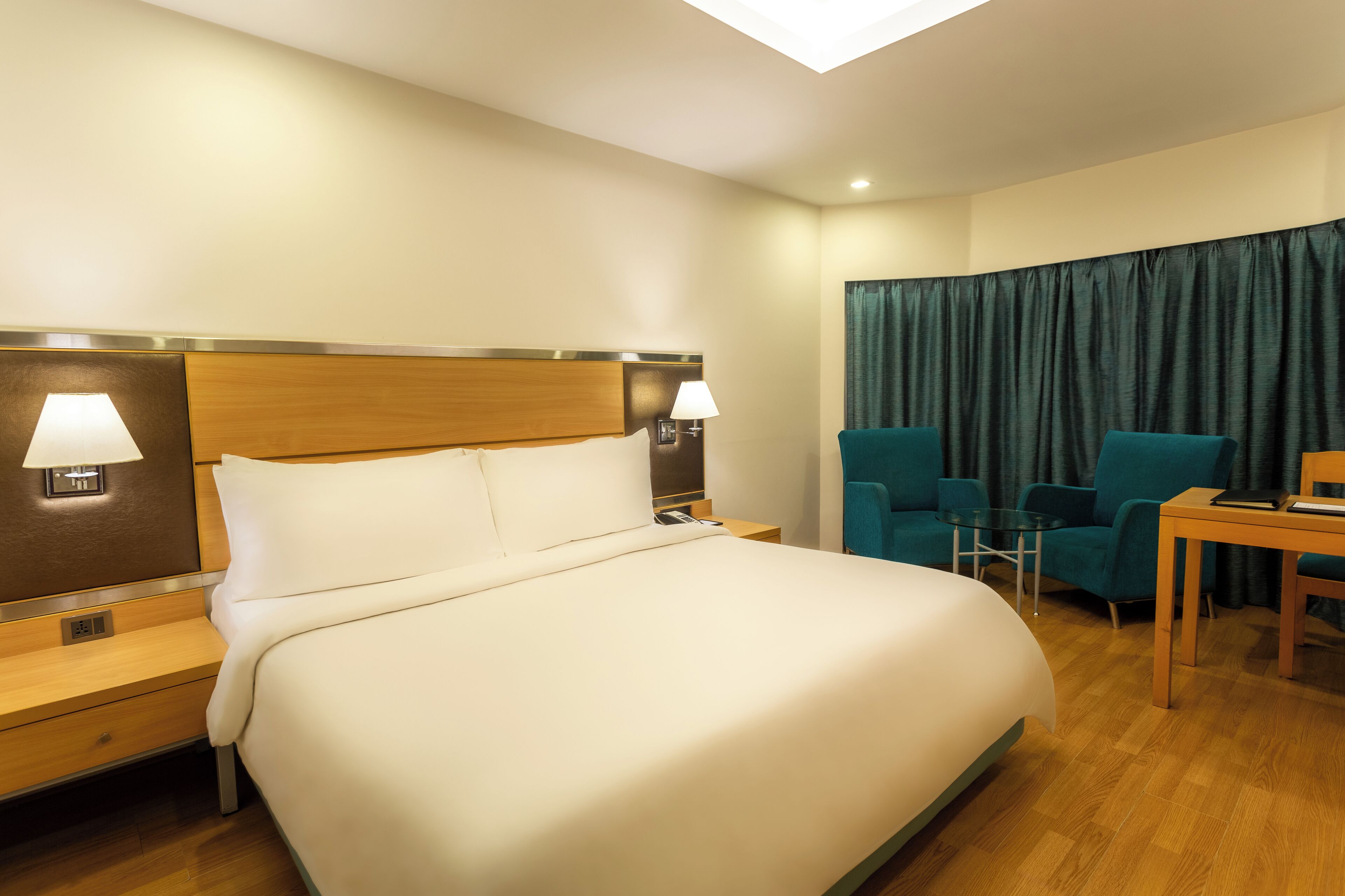 A hotel room with a large bed, two bedside tables with lamps, a desk with a chair, and a view of the city through a window with curtains.