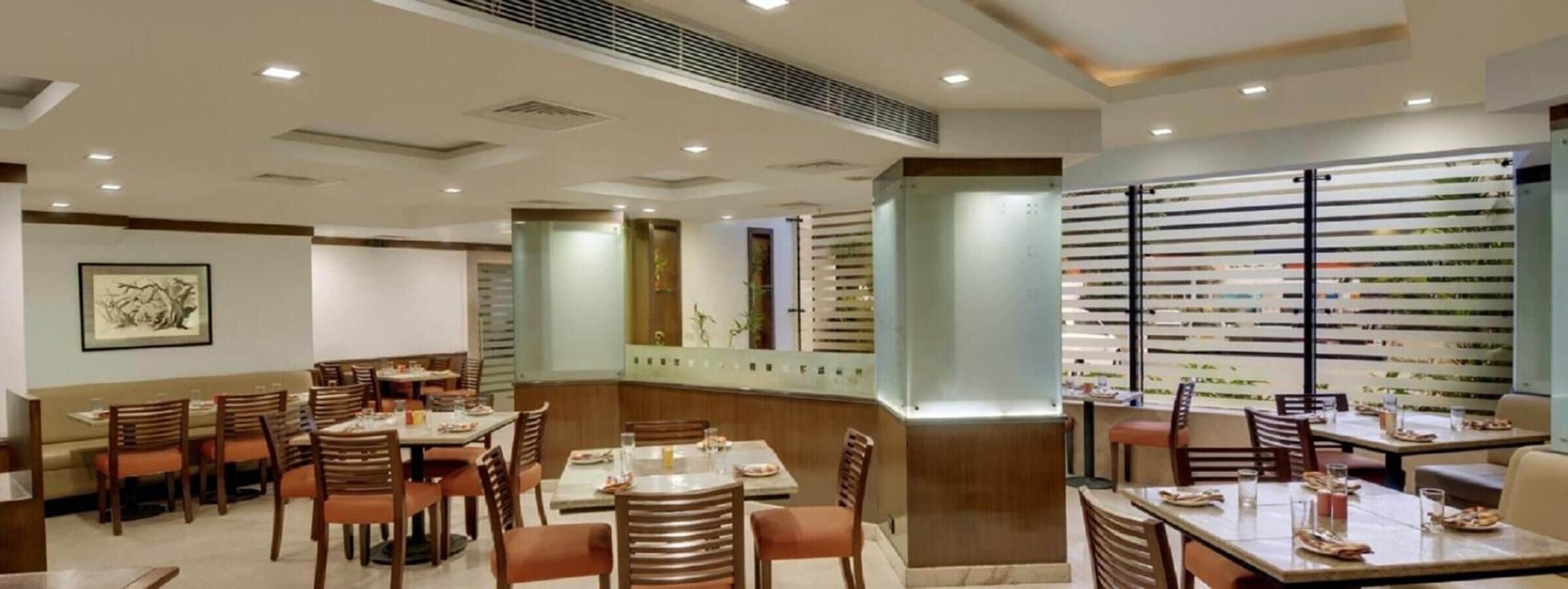 A restaurant with multiple dining tables and chairs, a large window with blinds, and a framed artwork on the wall.