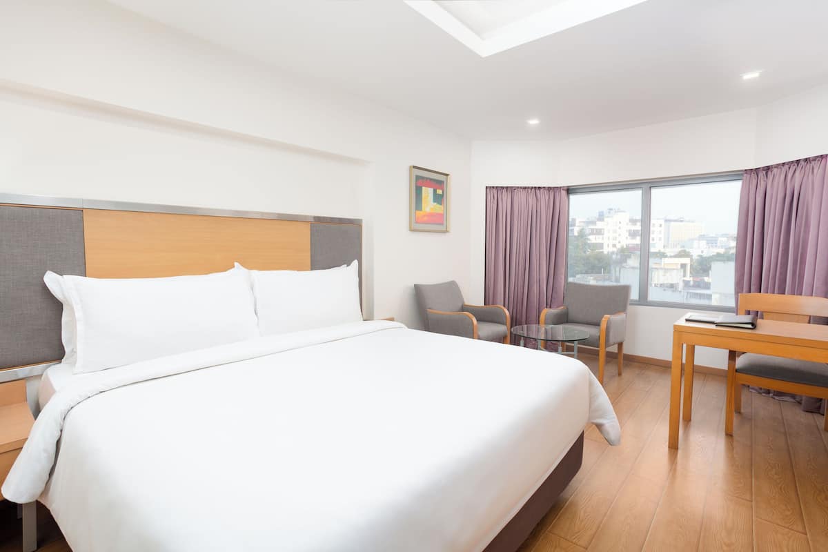 A hotel room with a large bed, a desk, a chair, and a view of the city through the window.