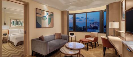 A modern hotel room with a bed, sofa, dining table, and chairs. There is a large window with a view of the city.