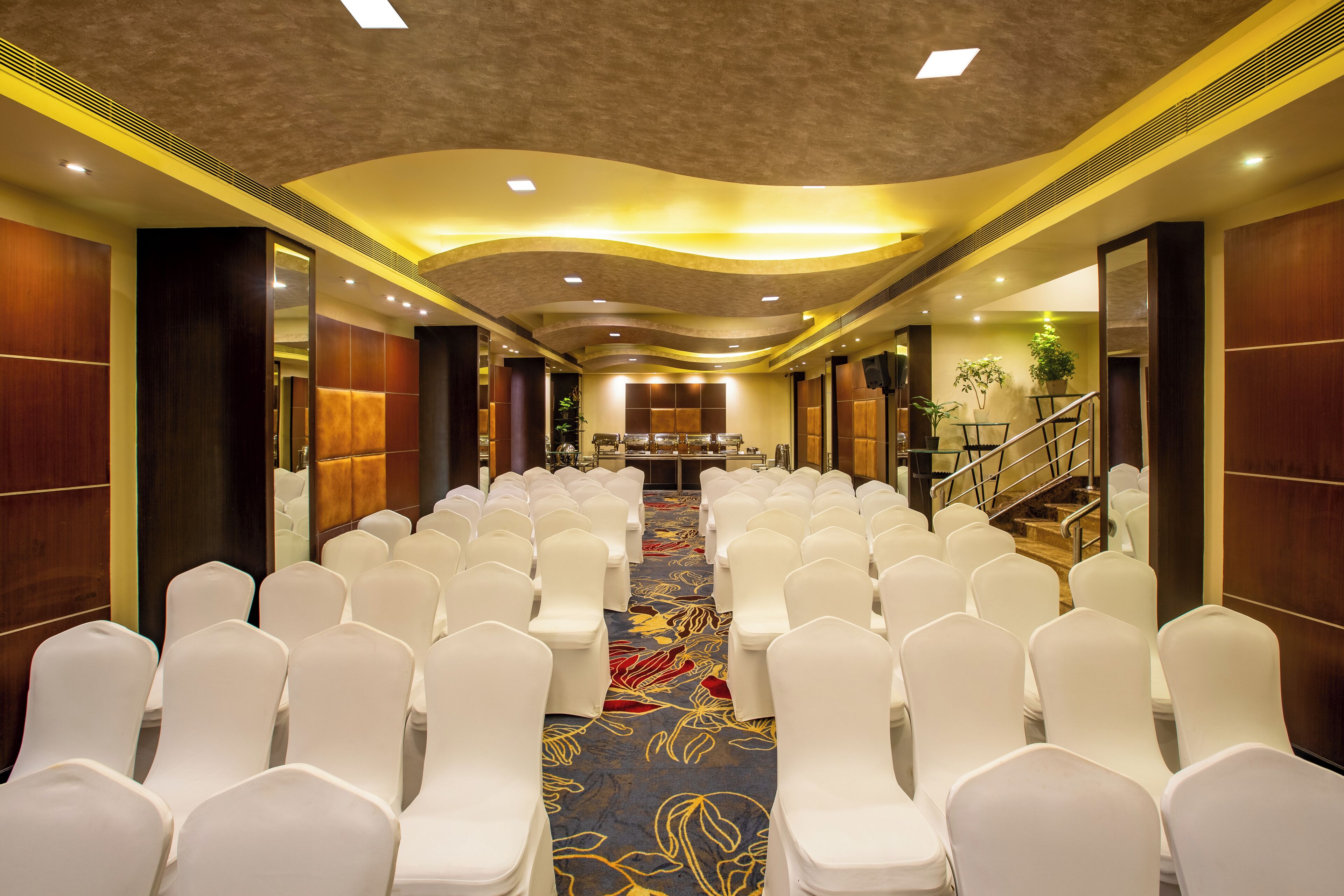Un salón de conferencias con filas de sillas blancas, suelo alfombrado y una escalera que sube a un nivel superior.