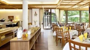 A restaurant with wooden tables and chairs, a buffet area, and large windows offering a view of the outdoors. - Europark Hotel International (Fuessen)