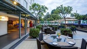 Un área de comedor al aire libre con mesas y sillas redondas, rodeada de follaje y un edificio con un letrero que dice 