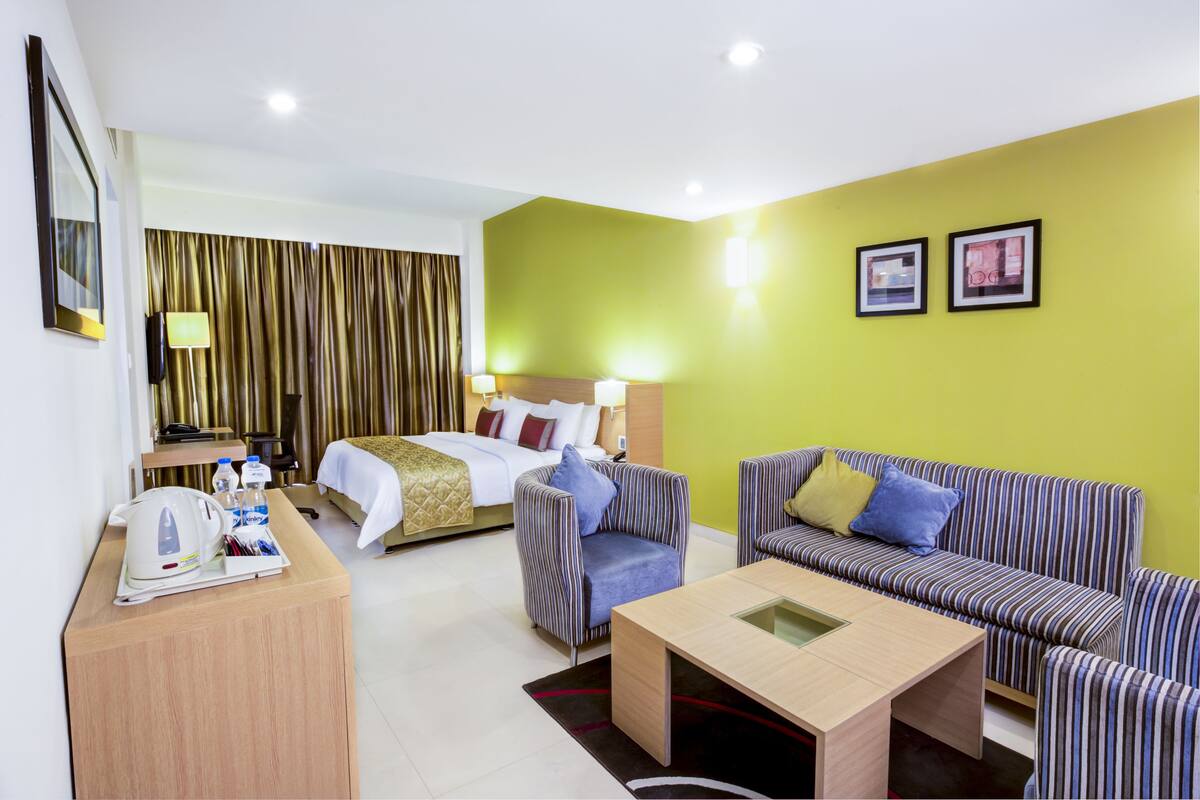 A hotel room with a bed, a sofa, a coffee table, a desk with a kettle and water bottles, and a wall with two framed pictures.