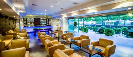 A modern lounge area with leather sofas and tables, a bar in the background, and a view of greenery outside.