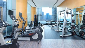 A modern gym with treadmills, ellipticals, and weight machines, large windows with a city view, and a mirrored wall.