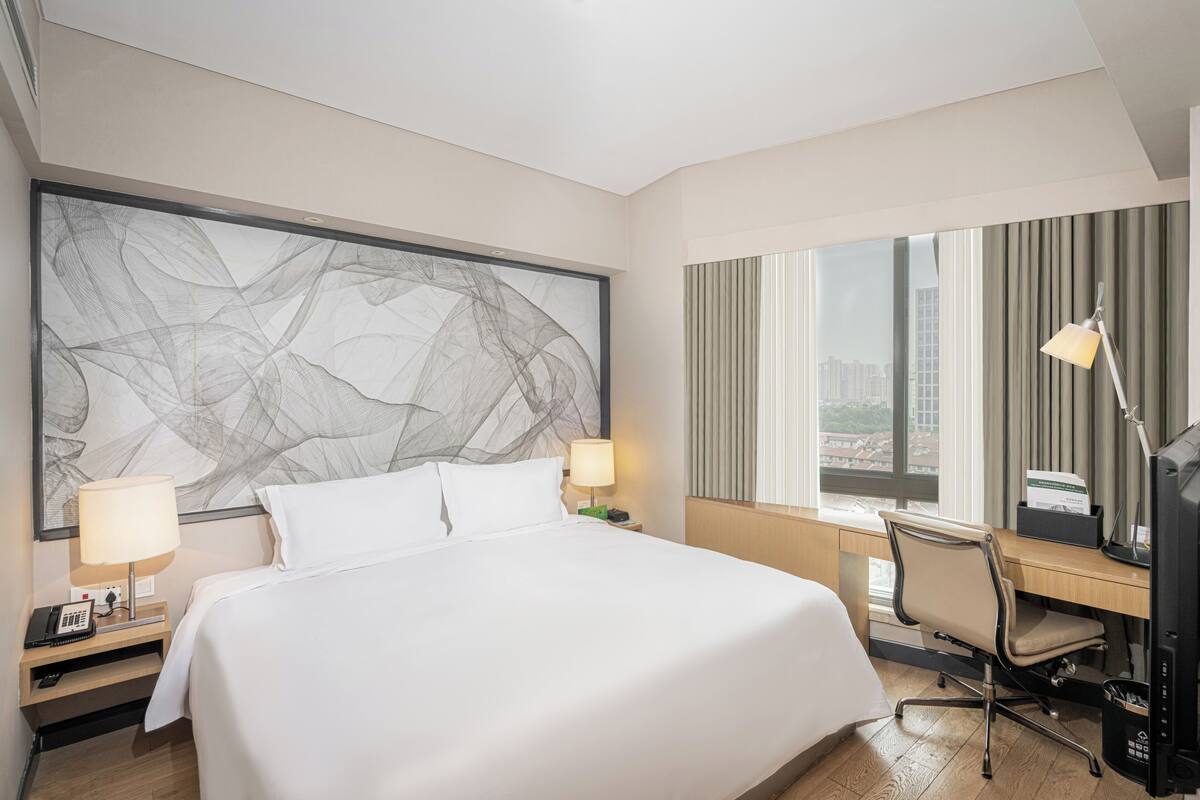 Een hotelkamer met een groot bed, twee nachtkastjes met lampen, een bureau met stoel, en een groot raam met gordijnen.