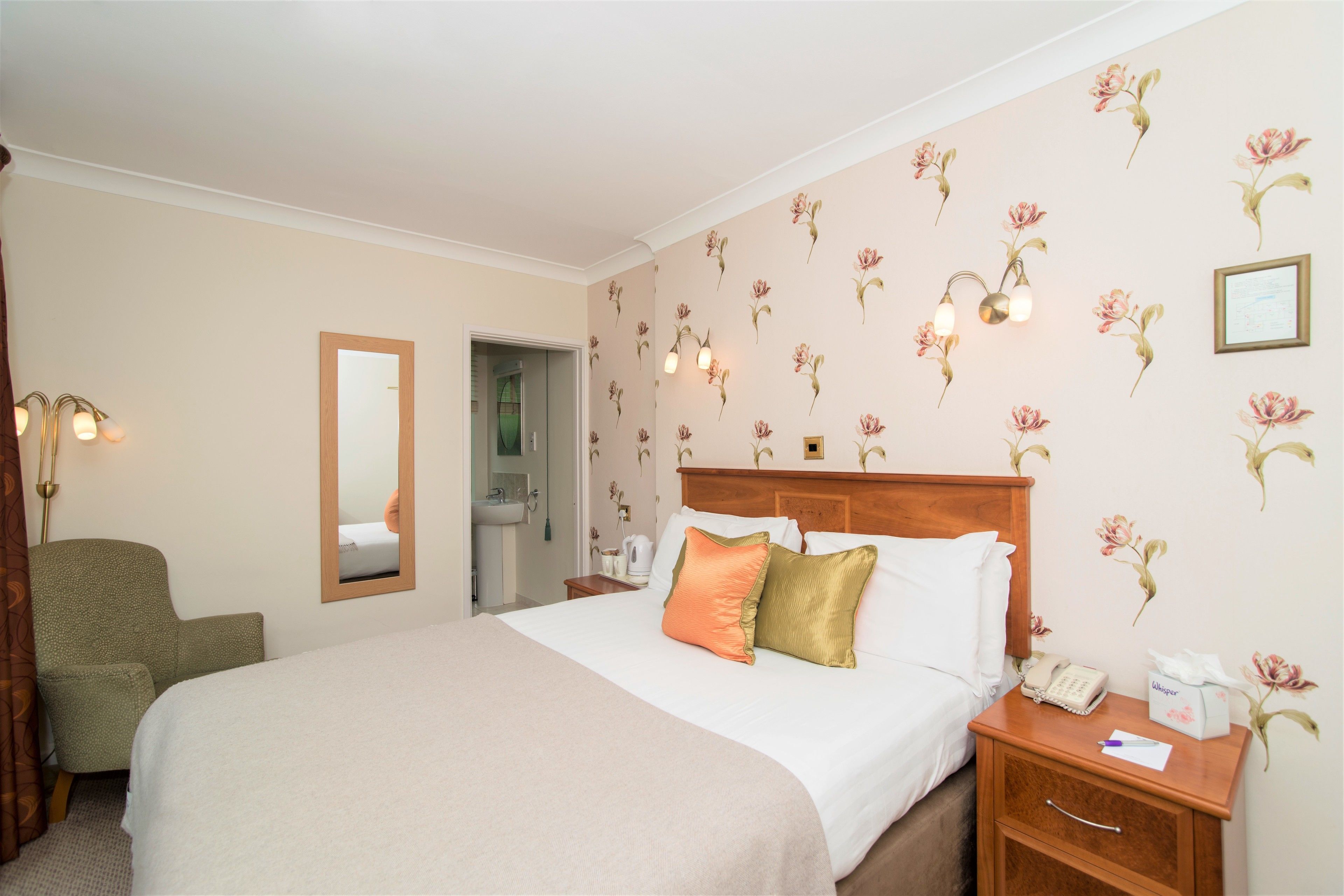 Een hotelkamer met een bed, een nachtkastje met telefoon en zakdoekjes, een stoel, een spiegel en een deur naar een badkamer.