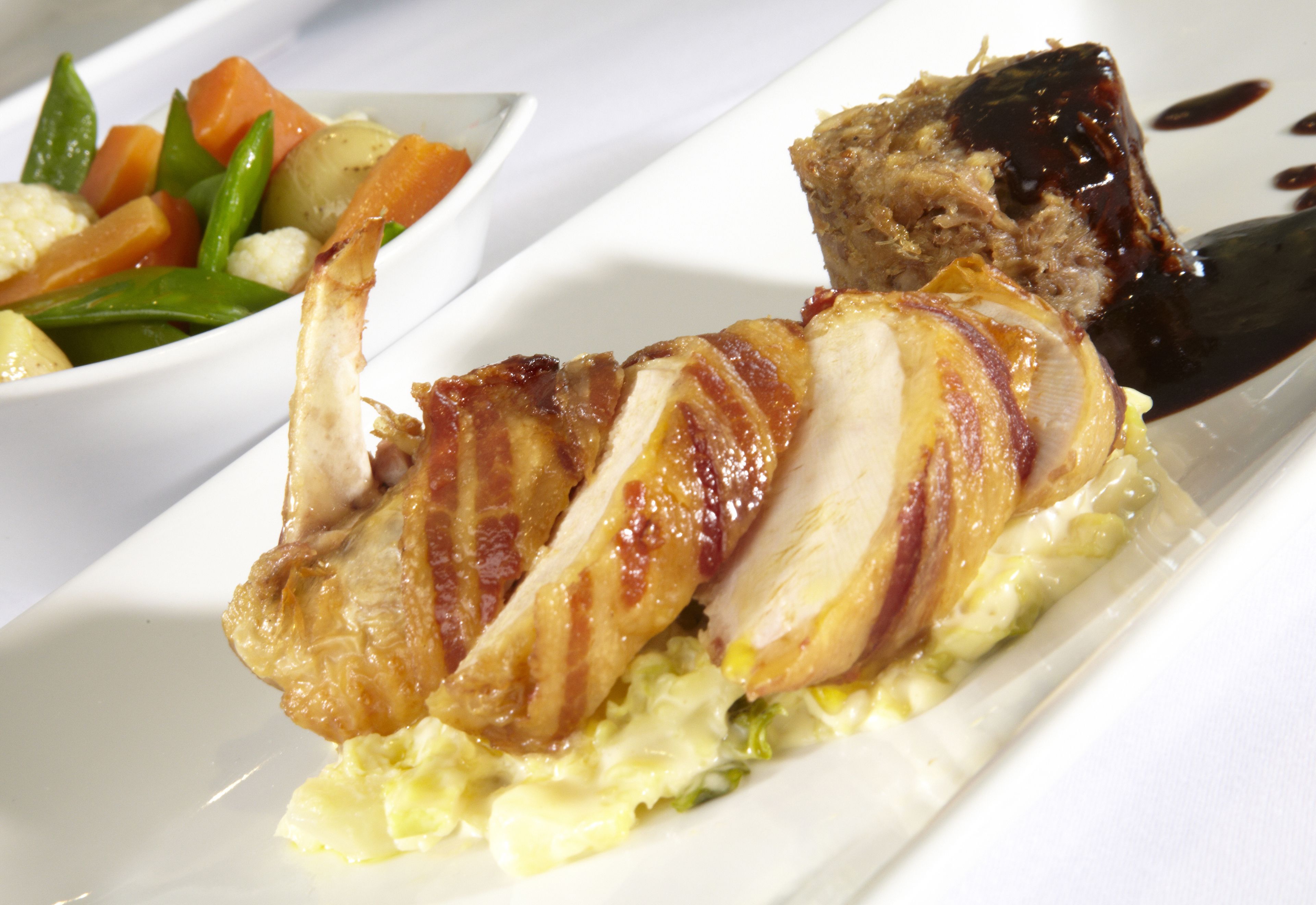 Un plato de comida con un trozo de carne envuelto en tocino, acompañamiento de verduras y salsa oscura.