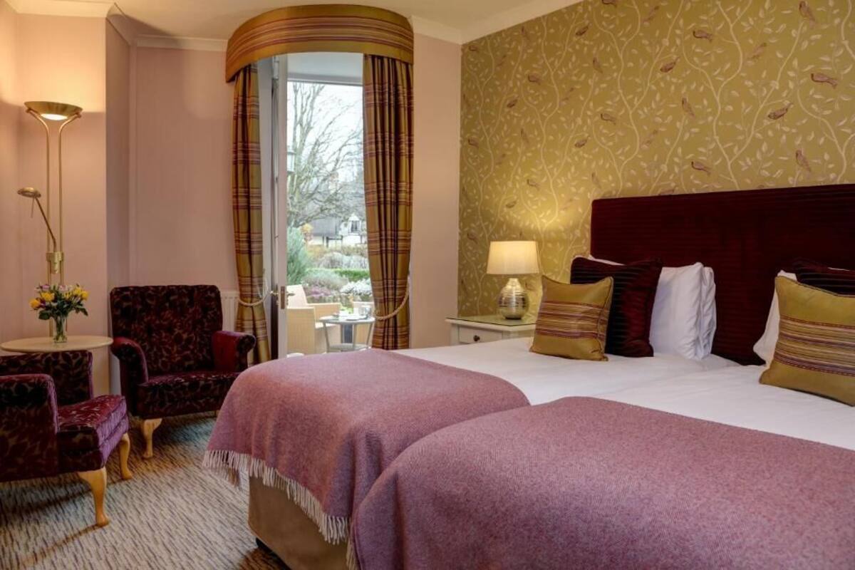 A hotel room with a large bed, two armchairs, a small table with a lamp, and a view of the outdoors.