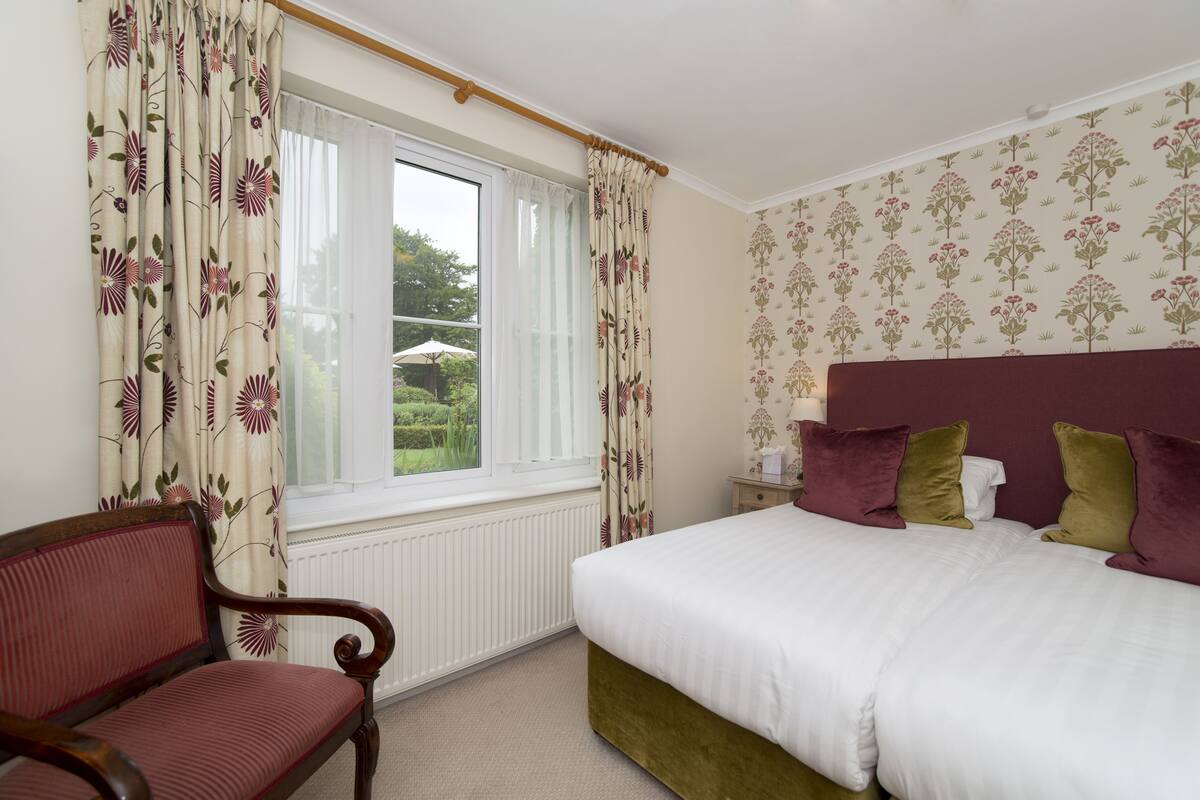 A bedroom with a bed, a chair, a window with curtains, and a wall with floral wallpaper.