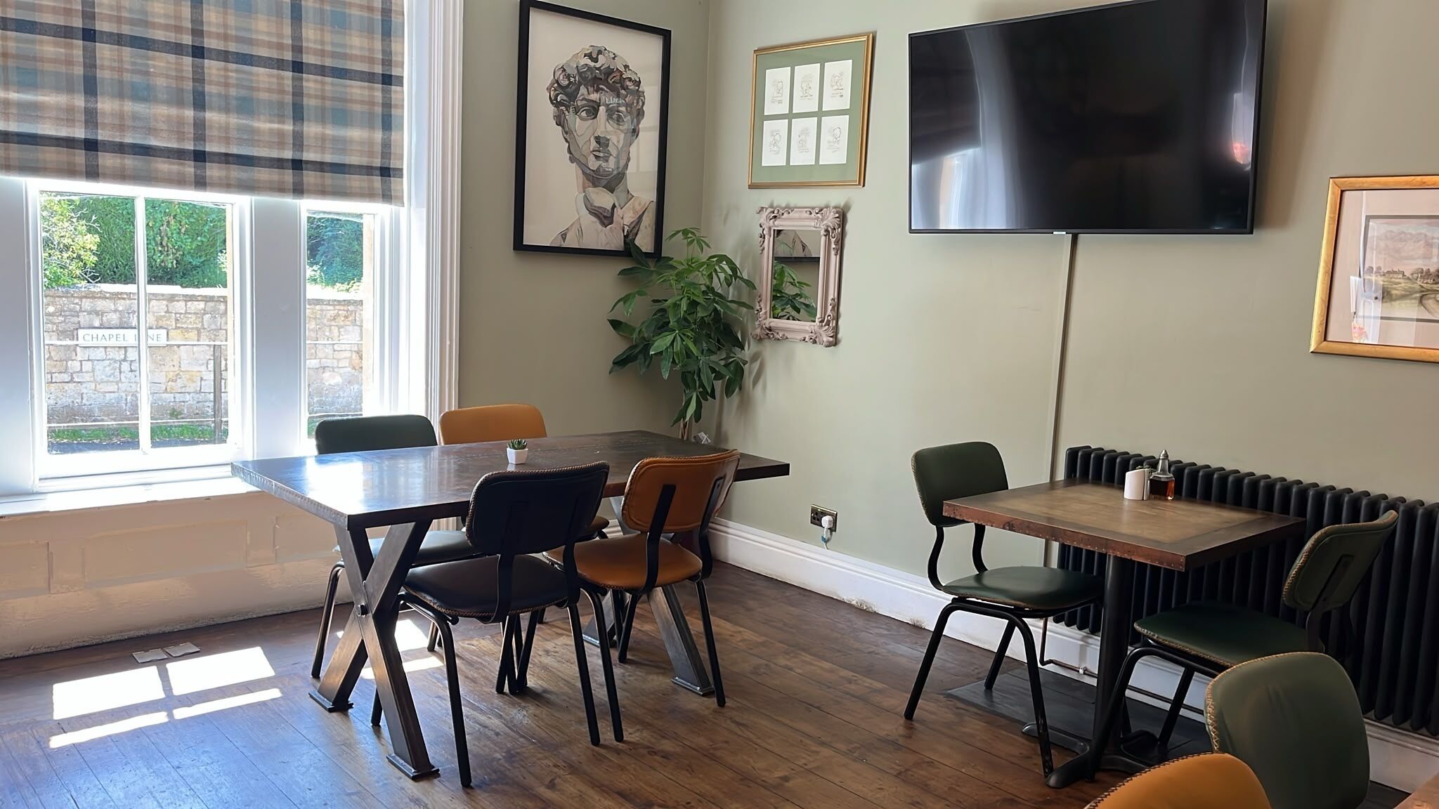 A dining area with a wooden table, chairs, a potted plant, a framed artwork, a television, and a window with a view of greenery.