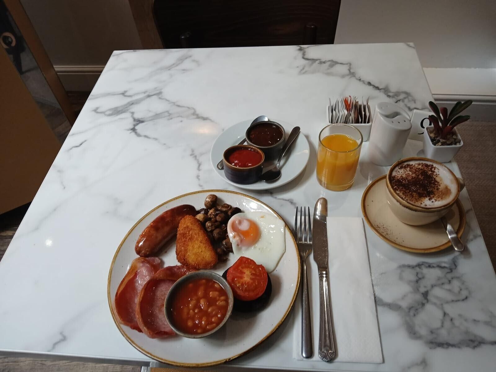 Ein Frühstückstisch mit Eiern, Würstchen, Bohnen und Tomaten, einer Kaffeetasse und einem Glas Orangensaft.