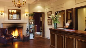 A lobby with a fireplace, leather armchair, wooden reception desk, and a chandelier. - Three Ways House Hotel (Chipping Campden)
