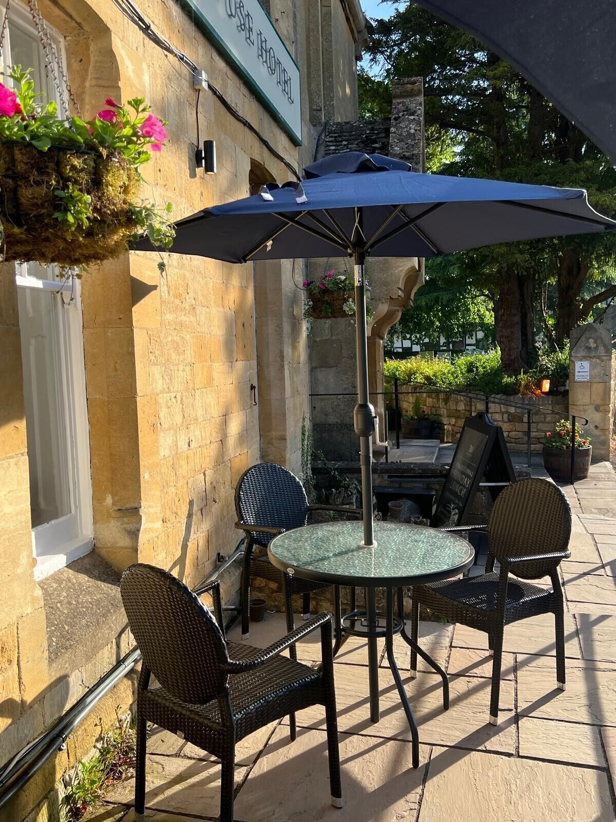 Un patio al aire libre con una mesa redonda y cuatro sillas de mimbre, una sombrilla y una cesta de flores colgante.