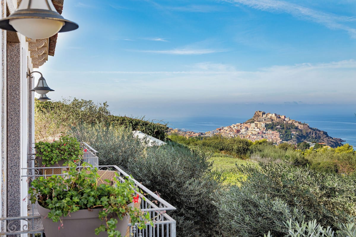 A balcony with potted plants, a view of a coastal town with buildings on a hill, and the sea in the background.