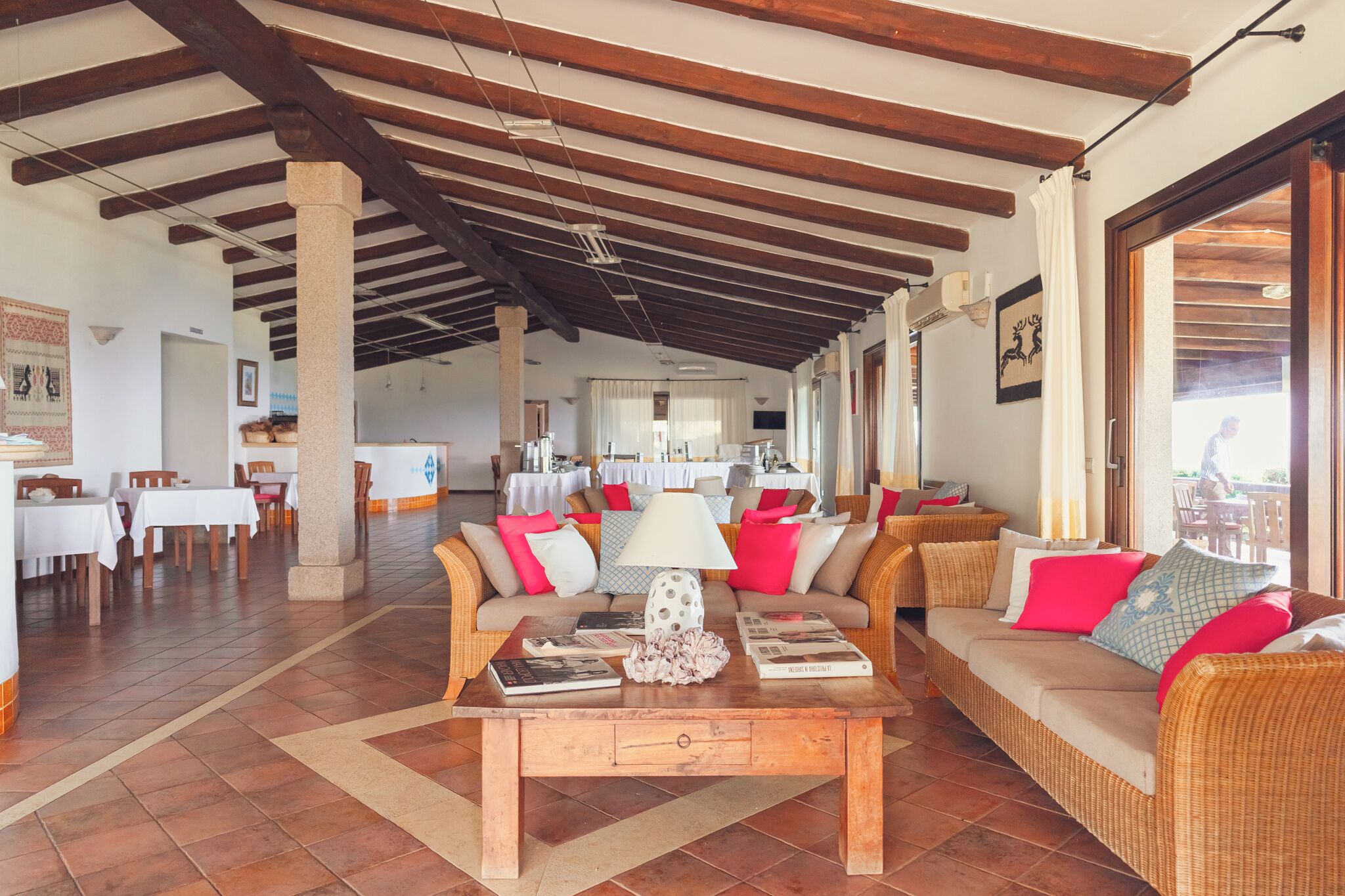 A spacious indoor area with a high ceiling, wooden beams, a central seating area with a table, and a dining area in the background.