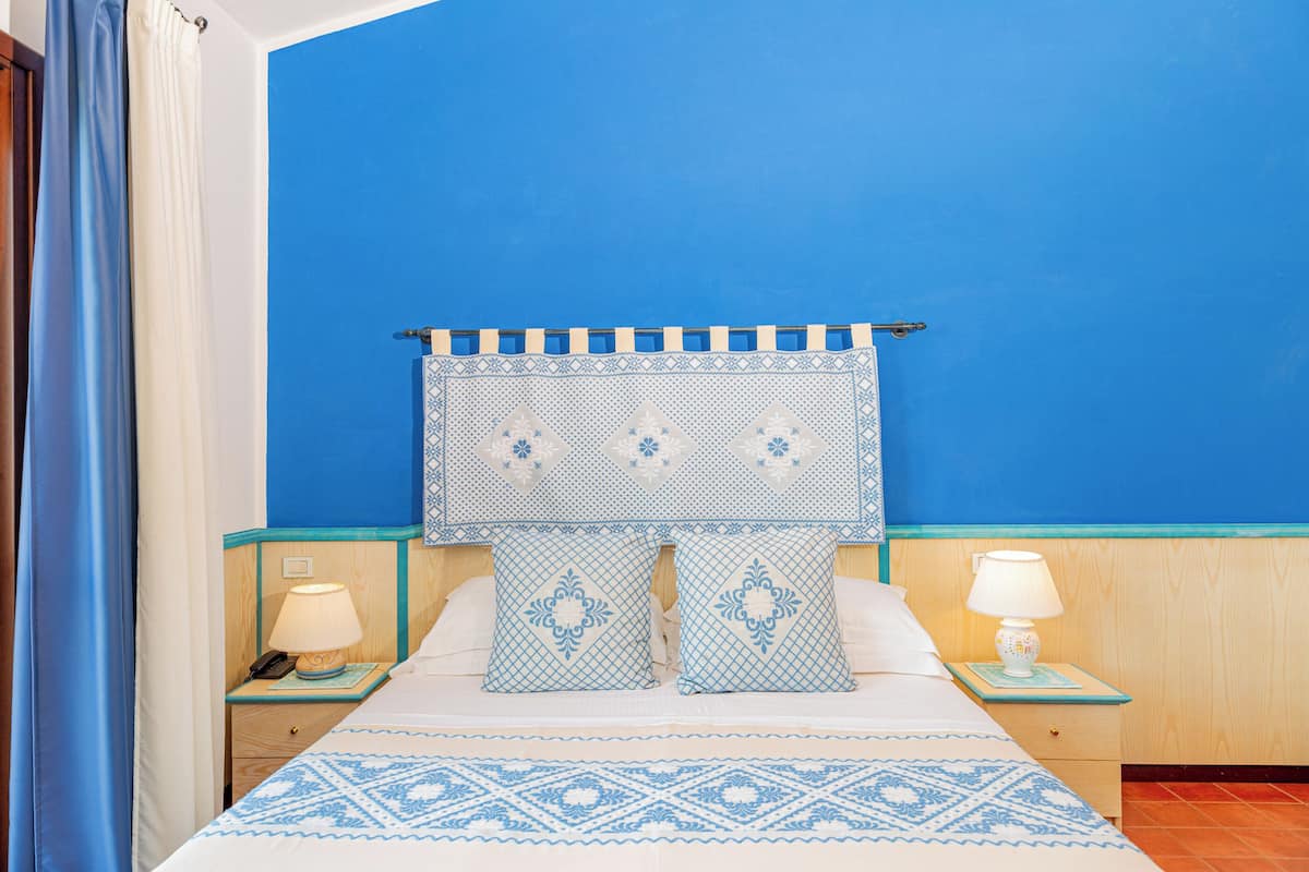 A room with a bed, two bedside tables, a nightstand, a headboard with a patterned curtain, and a blue wall.
