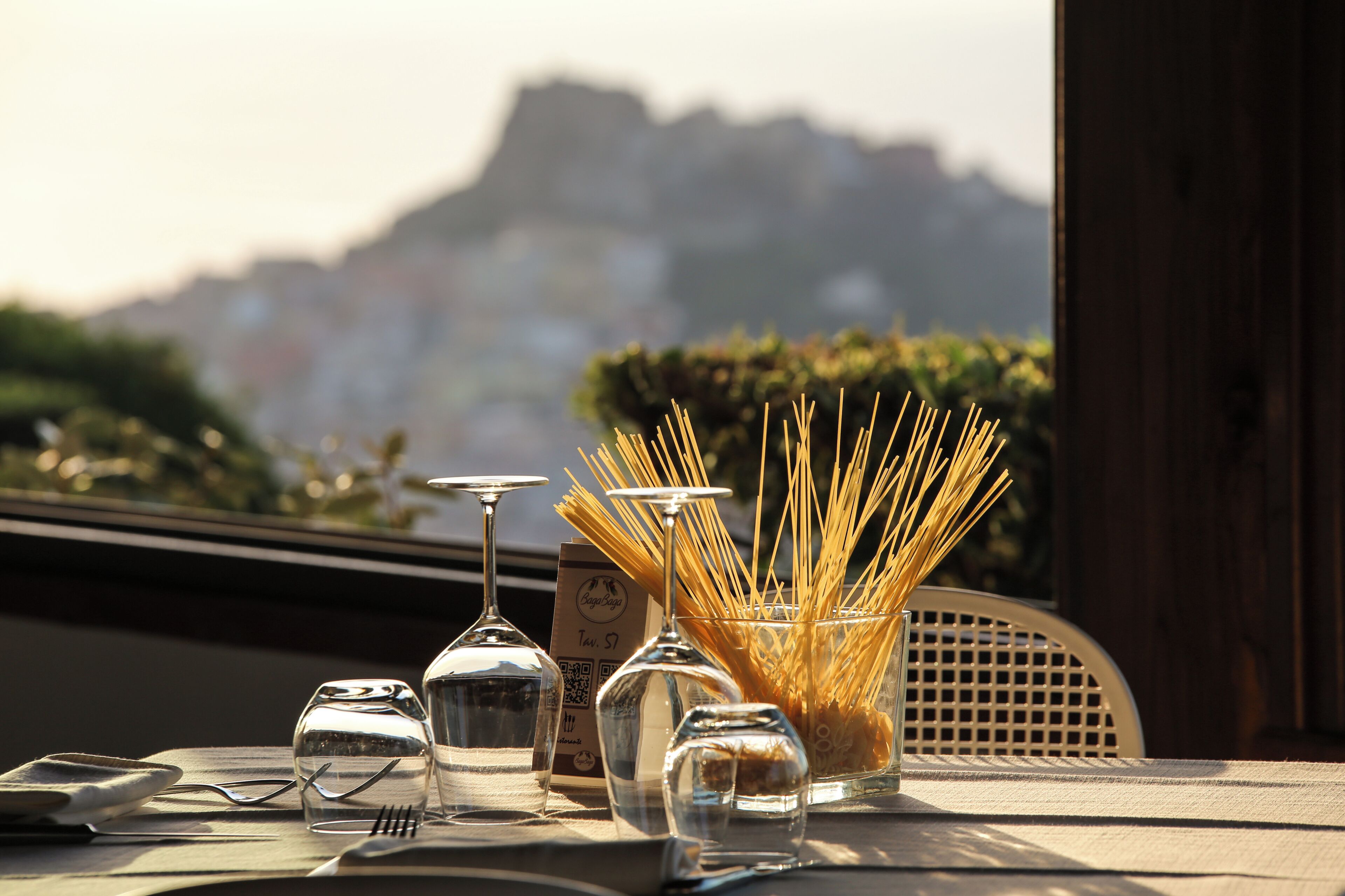 Una mesa dispuesta con copas de vino, cubiertos y un tazón de pasta, con vistas a un paisaje montañoso.