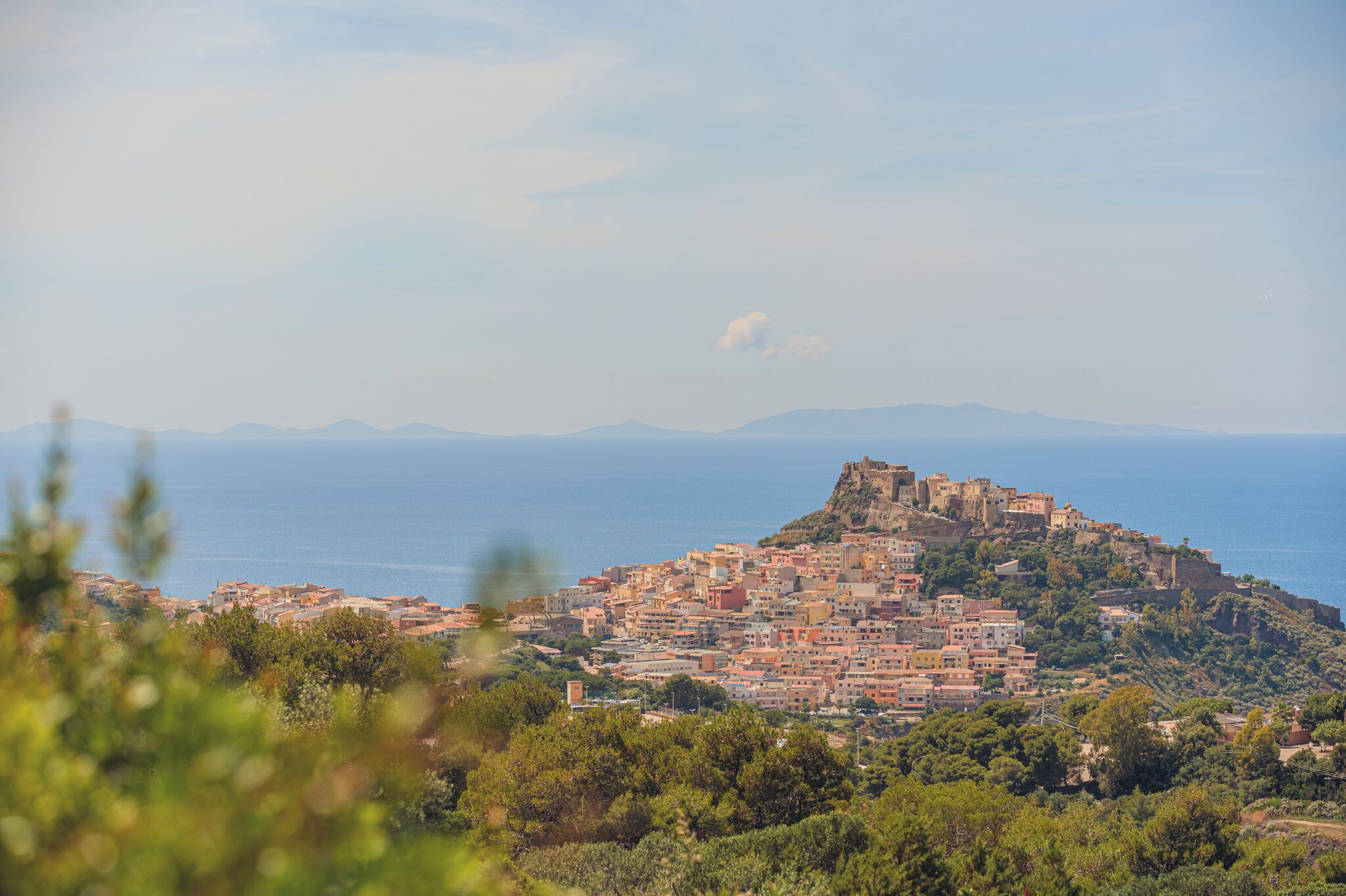 Uma cidade costeira com um castelo histórico numa colina, rodeada por edifícios e vegetação.