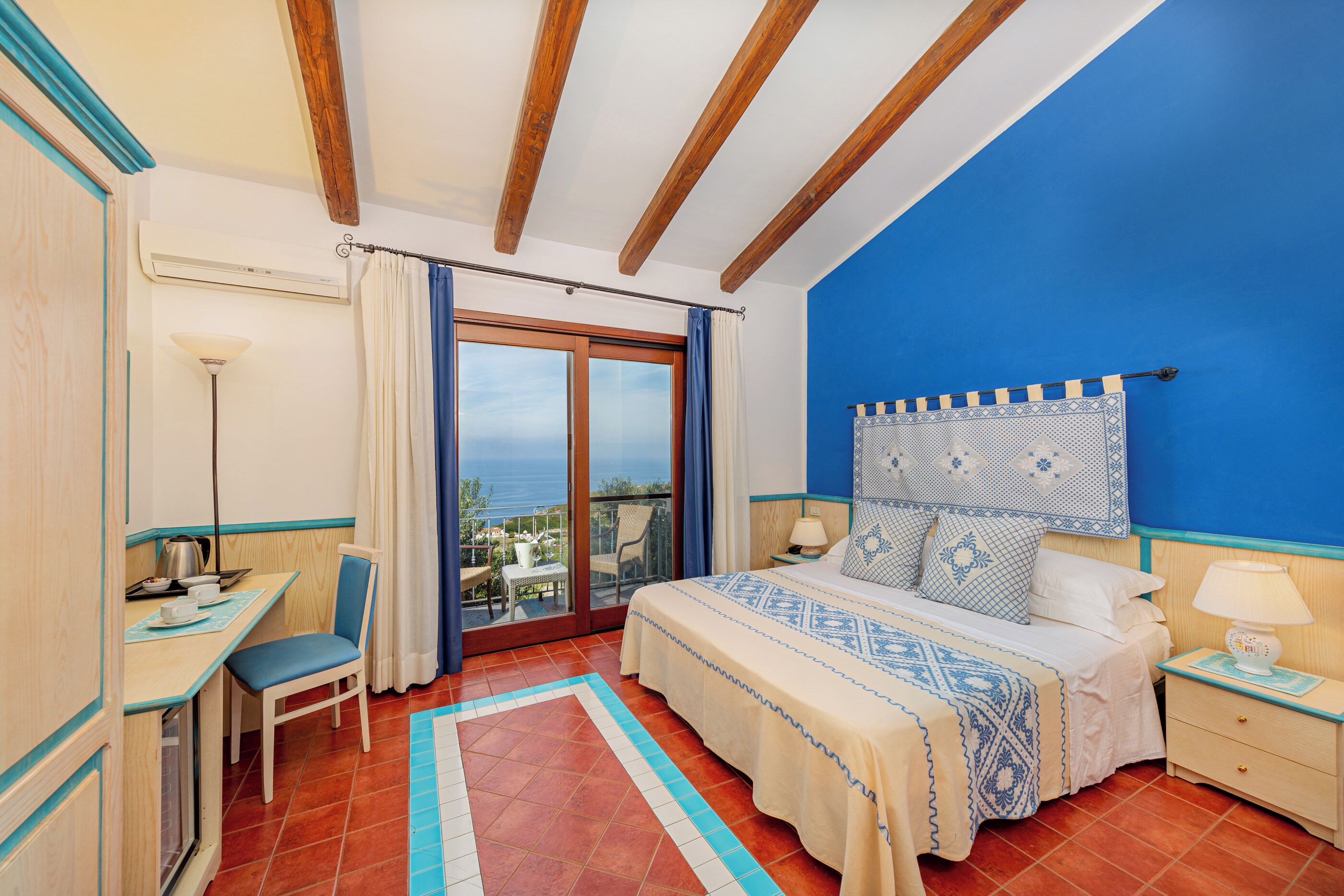 A bedroom with a bed, a desk, a chair, a nightstand, and a view of the sea.