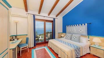 A bedroom with a bed, a desk, a chair, a nightstand, and a view of the sea.