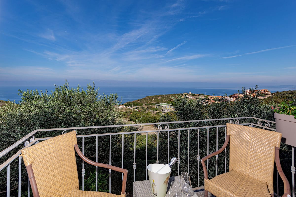 A balcony with wicker chairs, a table, and a view of the sea and distant buildings.